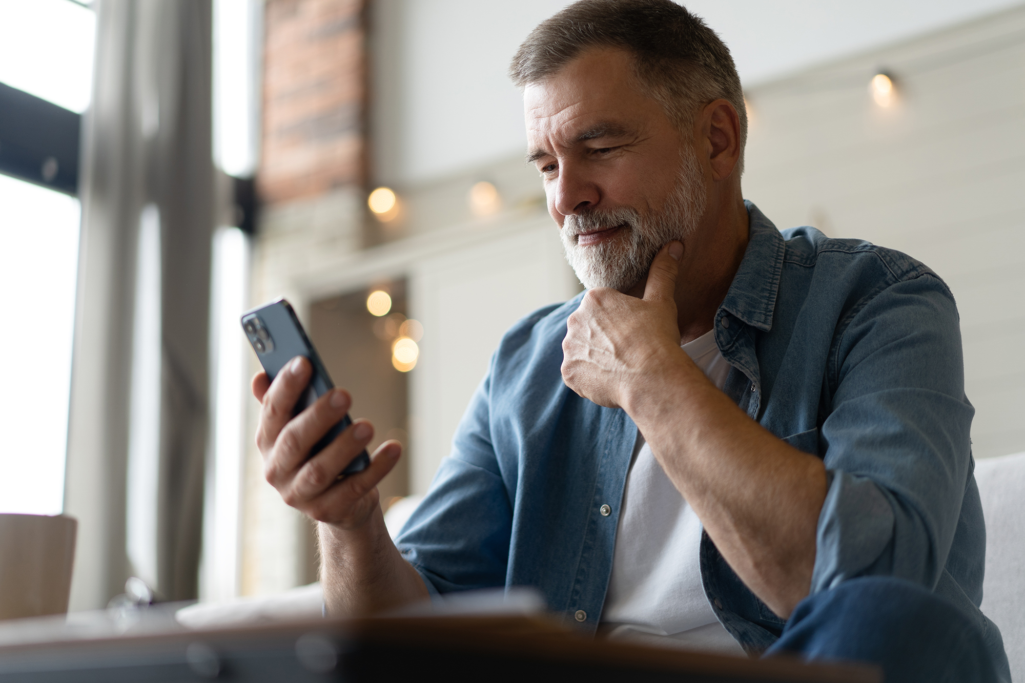 Happy smiling senior man using smartphone device to submit selfie pics of his smile to Tyler Glaser Dental while sitting on sofa at home. Mature man lying on couch reading messages on mobile phone, relaxing at home.
